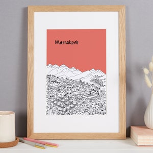 May include: A framed print of a black and white illustration of the city of Marrakech, Morocco. The illustration depicts a cityscape with mountains in the background. The print has a coral red background with the word "Marrakech" in black text.