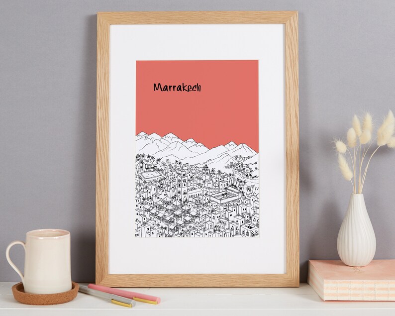 May include: A framed print of a black and white illustration of the city of Marrakech, Morocco. The illustration depicts a cityscape with mountains in the background. The print has a coral red background with the word "Marrakech" in black text.