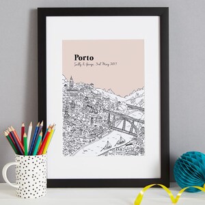 May include: Black framed print featuring a detailed black and white illustration of Porto, Portugal, with a pink background. The word "Porto" is at the top, with the names "Sally & George" and the date "3rd May 2017" below. A pencil cup and a blue paper decoration are in the foreground.