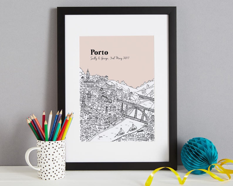 May include: Black framed print featuring a detailed black and white illustration of Porto, Portugal, with a pink background. The word "Porto" is at the top, with the names "Sally & George" and the date "3rd May 2017" below. A pencil cup and a blue paper decoration are in the foreground.