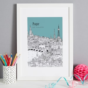 Op de afbeelding: Een zwart-witte illustratie van de skyline van Praag met de tekst "Prague Girls Weekend 2016" op een teal achtergrond. De illustratie is ingelijst in een wit frame.