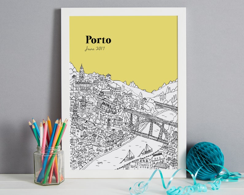 May include: A framed black and white illustration of Porto, Portugal, with a yellow background. The artwork depicts the city's architecture, a bridge, and the text "Porto June 2017". Colored pencils in a glass jar and a blue paper decoration are in the foreground.