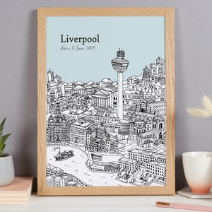 Op de afbeelding: Een zwart-witte lijntekening van de skyline van Liverpool, inclusief het Liver Building, het Royal Liver Building en de Radio City Tower. De tekening is ingelijst in een licht houten lijst en heeft een lichtblauwe achtergrond. De tekst "Liverpool Katie & Sam 2015" is onder de tekening gedrukt.