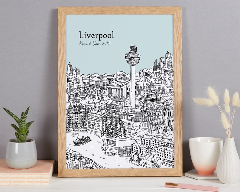 Op de afbeelding: Een zwart-witte lijntekening van de skyline van Liverpool, inclusief het Liver Building, het Royal Liver Building en de Radio City Tower. De tekening is ingelijst in een licht houten lijst en heeft een lichtblauwe achtergrond. De tekst "Liverpool Katie & Sam 2015" is onder de tekening gedrukt.