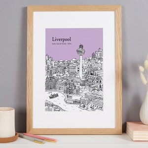Op de afbeelding: Een ingelijste print van een zwart-witte lijntekening van de skyline van Liverpool. De tekening omvat het Liver Building, het Royal Liver Building en de Radio City Tower. De achtergrond is lichtpaars. De print is getiteld "Liverpool" en bevat de tekst "Jude, Sam & Verity - 2014".