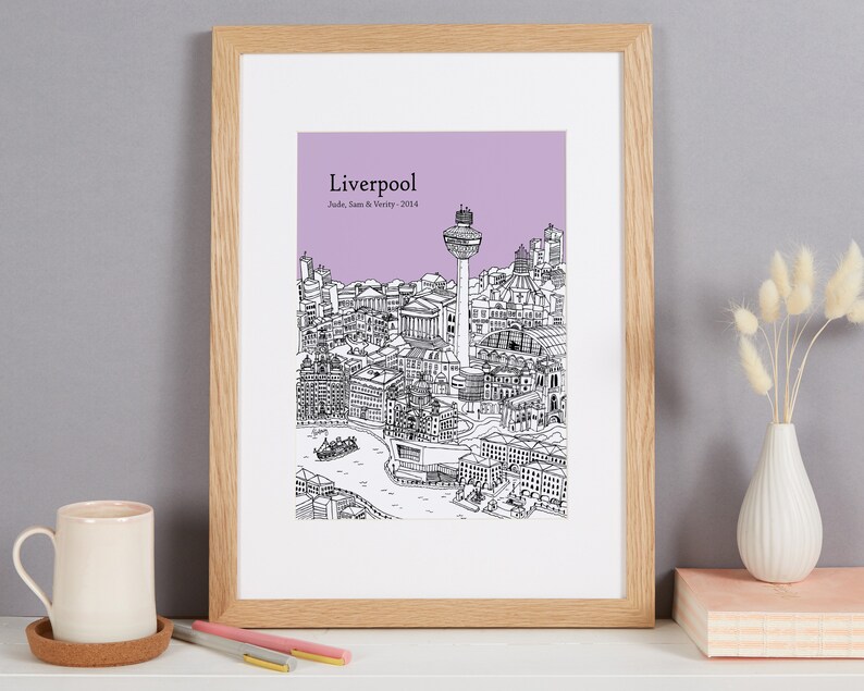 Op de afbeelding: Een ingelijste print van een zwart-witte lijntekening van de skyline van Liverpool. De tekening omvat het Liver Building, het Royal Liver Building en de Radio City Tower. De achtergrond is lichtpaars. De print is getiteld "Liverpool" en bevat de tekst "Jude, Sam & Verity - 2014".