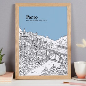 May include: A framed art print featuring a detailed black and white illustration of Porto, Portugal, with a blue sky background. The text "Porto" and "Our first holiday, May 2018" are printed at the top. The frame is made of light-colored wood.