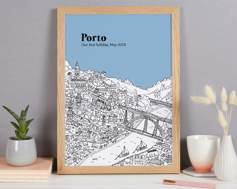 May include: A framed art print featuring a detailed black and white illustration of Porto, Portugal, with a blue sky background. The text "Porto" and "Our first holiday, May 2018" are printed at the top. The frame is made of light-colored wood.