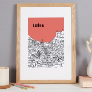 May include: A framed print of a black and white illustration of the city of Lisbon, Portugal. The illustration is set against a coral pink background. The word "Lisbon" is printed in black at the top of the illustration.