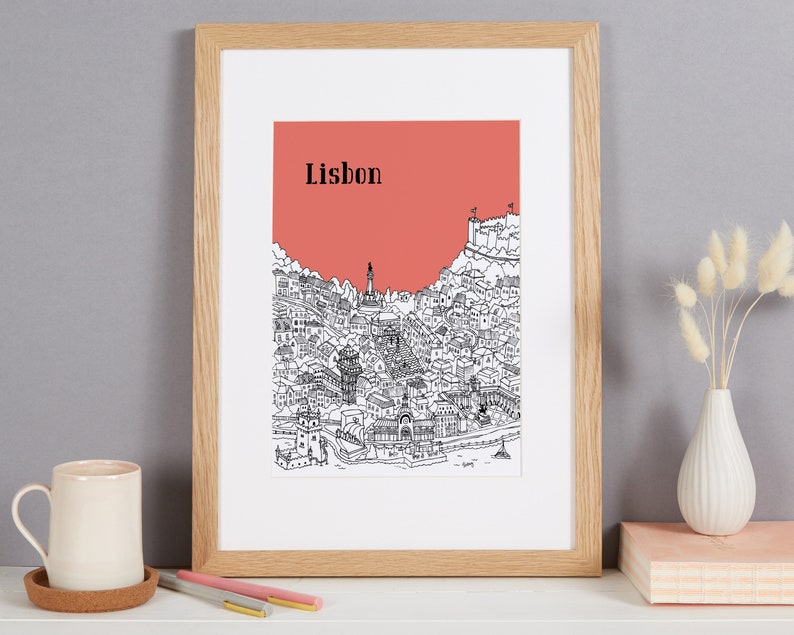 May include: A framed print of a black and white illustration of the city of Lisbon, Portugal. The illustration is set against a coral pink background. The word "Lisbon" is printed in black at the top of the illustration.