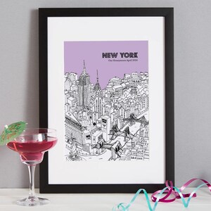 May include: Black framed print of a New York City skyline in black and white, with a purple background. The text "NEW YORK" and "Our Honeymoon April 2016" are visible. A cocktail with a green umbrella is in the foreground.