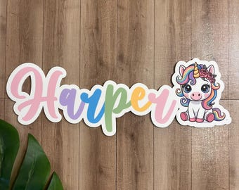 May include: A wooden cutout spelling "Harper" in a rainbow gradient with a white border. A cartoon unicorn with a rainbow mane and flower crown is to the right. The background is a wood grain pattern.