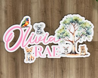 May include: A white wooden sign with the names Olivia Rae in pink cursive lettering. The sign is decorated with a watercolour painting of a tree, a koala, a bird, and two other animals.