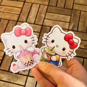 May include: Two die-cut stickers featuring the character Hello Kitty. One sticker shows Hello Kitty holding a pink drink with boba, and the other holds a green drink. Both stickers have a white background and a pink bow.