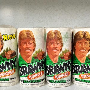 May include: Four rolls of Brawny paper towels, each featuring a different design. The rolls are white with green and black text and a picture of a man with brown hair and a green shirt. The text on the rolls reads "Brawny Designer", "Brawny Prints", "Brawny Prints", and "Brawny Designer".