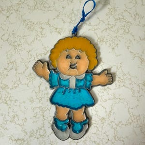 May include: A vintage-style ornament featuring a cartoon doll with yellow hair and a blue dress. The doll has outstretched arms and is outlined in silver. A blue string is attached for hanging.