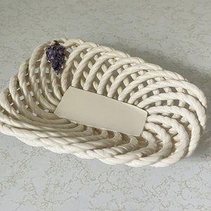 May include: White ceramic bowl with a woven rope design. The bowl has a small purple grape design on the side.