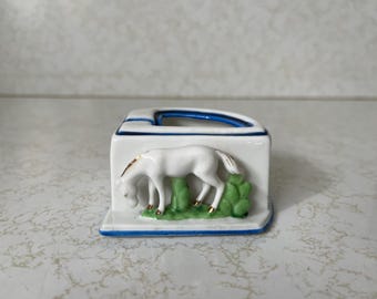 May include: A white ceramic desk organizer with a blue and gold trim. The organizer features a white horse grazing on green grass. The top has a blue curved opening. A decorative piece for a desk or office.