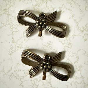 May include: Two antique-style metal bow-shaped ornaments. Each bow has a central cluster of small, round details and a loop at the top. The metal has a tarnished, aged appearance, suggesting vintage or antique origins. The background is a light, speckled surface.