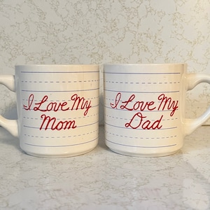 May include: Two off-white ceramic mugs with curved handles. Each mug has a lined paper design with the words "I Love My Mom" and "I Love My Dad" in red script. A heartwarming gift for parents.