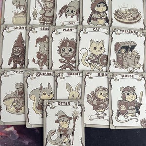 May include: A selection of illustrated fantasy cards depicting animals and creatures. The cards feature designs such as a bear fishing, a spider, a hamster, and a treasure chest. The cards are in a neutral colour palette.