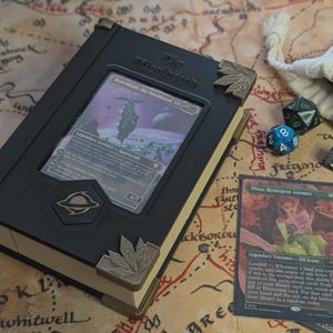 Könnte beinhalten: Eine schwarze, buchförmige Kartenaufbewahrungsbox mit metallischen Eckakzenten und einem Sichtfenster, das eine Hearth-hull, the Worldseed Karte zeigt. Die Box hat ein kreisförmiges Emblem und wird von Spielwürfeln und Karten begleitet.