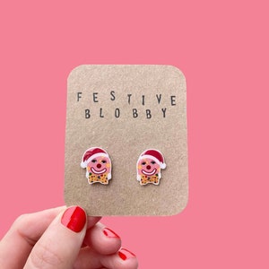 May include: A pair of festive clown earrings on a brown card, held by a hand with red nail polish. The earrings feature a clown face with a Santa hat and bow tie. The card reads "FESTIVE BLOBBY" in black letters.