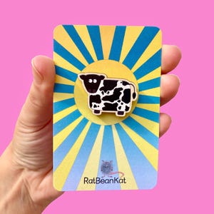Puede incluir: Un pin con forma de vaca en blanco y negro sobre una tarjeta amarilla y azul con diseño de rayos de sol. La tarjeta tiene el texto "RatBeanKat" en la parte inferior. El fondo es de color rosa liso.