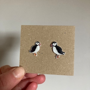 May include: A pair of puffin bird earrings. Each earring features a black and white puffin with a red beak and orange feet. The earrings are displayed on a brown square card, held by a hand.