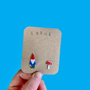 May include: A pair of earrings featuring a gnome and a mushroom. The gnome is wearing a red hat, blue shirt, and brown trousers. The mushroom is red with white spots. The earrings are on a brown card with the word "GNOME" printed on it.