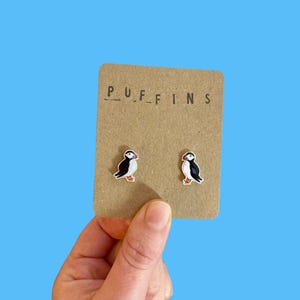 以下が含まれることがあります： 茶色のカードに、ツノメドリのイヤリングが2つ。イヤリングは黒、白、オレンジ色のツノメドリのデザインです。カードには「PUFFINS」の文字が印刷されています。背景は明るい青色です。