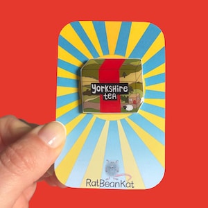 Peut inclure: Une broche émaillée rectangulaire illustrant un paysage vert avec une bande verticale rouge et le texte Yorkshire tea. La broche est fixée sur une carte de présentation à rayons jaunes et bleus avec un petit rat et le texte RatBeanKat.