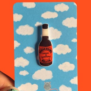 Pode incluir: Pin com uma garrafa castanha de Henderson's Relish sobre um fundo azul com nuvens brancas. A garrafa tem um rótulo vermelho com texto branco. O fundo tem o texto "RatBeanKat" e um rato de desenho animado.