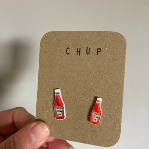 May include: A pair of novelty earrings shaped like miniature ketchup bottles. The earrings are red with white labels and feature the text "Heinz Tomato Ketchup". They are mounted on a brown card with the word "CHUP" printed at the top.