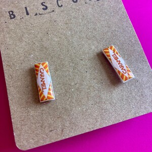 May include: A pair of caramel-themed earrings. Each earring is a rectangular shape with a white center and orange and yellow stripes. The word "CARAMEL" is printed on each earring. The earrings are on a brown card with the word "BISCUITS" printed at the top.