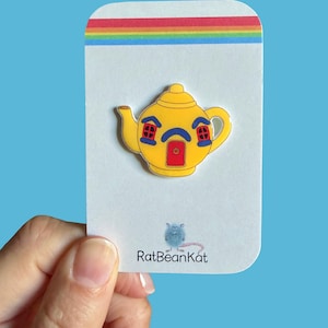 May include: A yellow teapot-shaped enamel pin featuring a red door, two blue-framed windows, and a blue arched brow design. The pin is attached to a white display card with a rainbow stripe at the top and the text RatBeanKat with a blue rat icon.