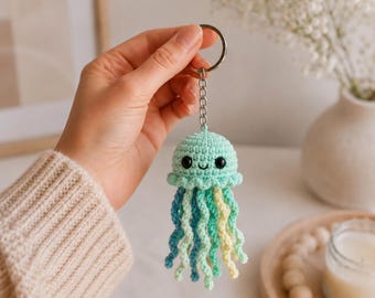 Könnte beinhalten: Ein gehäkelter Quallen-Schlüsselanhänger mit einem mintgrünen Kopf, schwarzen Perlenaugen und einem kleinen gestickten Lächeln. Die Qualle hat mehrere herabhängende, spiralförmige Tentakel in Blau-, Türkis-, Mintgrün- und Hellgelbtönen an einem Metallring.