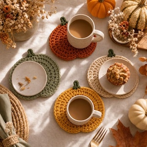 May include: Three round crochet coasters shaped like pumpkins in orange, green, and yellow, each featuring a small dark green stem. Two coasters hold ceramic mugs filled with coffee, while the third holds a small plate with a muffin.