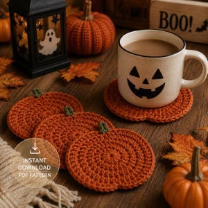 May include: Three round, orange, crocheted pumpkin-shaped coasters with small green stems. One coaster sits under a white ceramic mug featuring a black jack-o'-lantern face. The scene includes a black lantern, a small orange pumpkin, and autumn leaves.