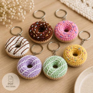 Peut inclure: Un ensemble de six porte-clés en forme de beignet crocheté dans des couleurs variées, notamment marron, rose, jaune, blanc, violet et vert. Chaque beignet est doté d'un anneau métallique et décoré de vermicelles en laine ou de glaçage.