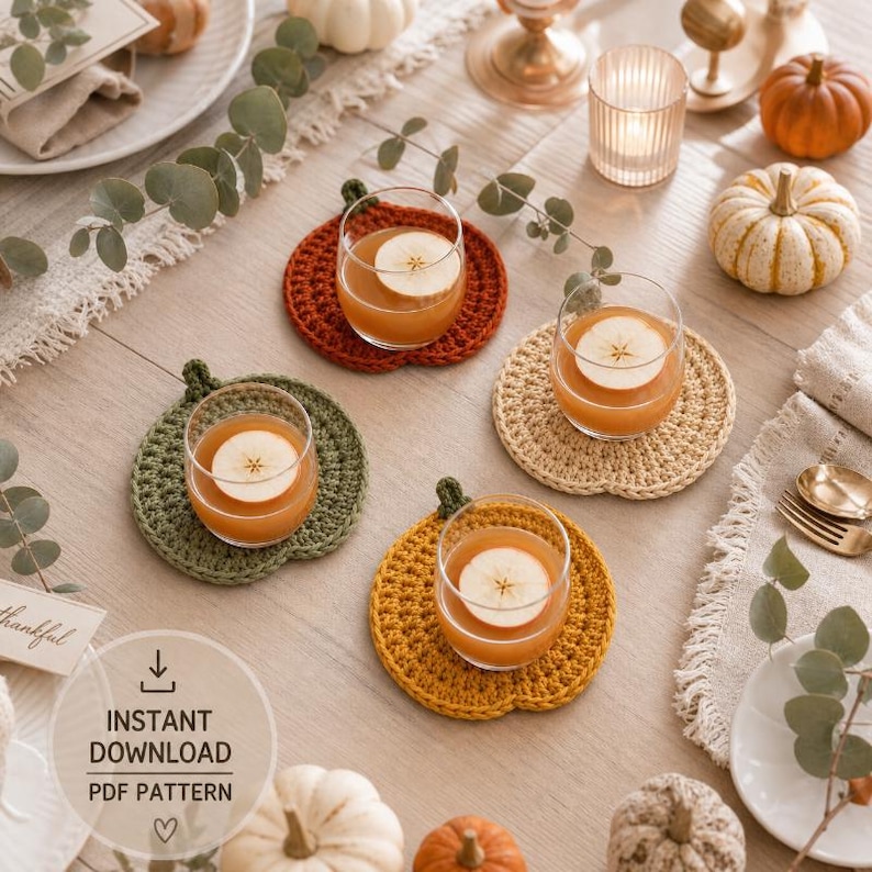 May include: Four round, pumpkin-shaped crochet coasters in rust, olive green, beige, and mustard yellow. Each coaster holds a clear glass filled with amber liquid and a thin apple slice. The coasters feature a small stem detail at the top.