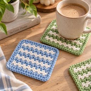 May include: Three square crochet coasters featuring a textured stitch pattern. One coaster is light blue with horizontal white stripes, while two coasters are sage green with horizontal white stripes. Each coaster has a thick, durable yarn construction.