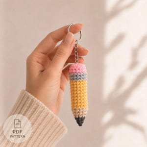 May include: A crochet pencil keychain featuring a pink eraser top, a grey metal band, a yellow pencil body, a tan wood-colored section, and a dark grey pointed tip. The keychain is attached to a silver-colored metal ring with a short chain link.