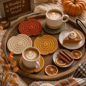 May include: A set of three round, crocheted pumpkin-shaped coasters in orange, yellow, and cream colors. The coasters are arranged on a wooden tray alongside two ceramic mugs filled with latte art, a slice of pumpkin pie, and cinnamon sticks.
