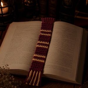 May include: A rectangular crochet bookmark featuring a deep maroon base with horizontal stripes in a golden yellow yarn. The bookmark has a textured stitch pattern and is finished with a fringe of golden yellow yarn at the bottom edge.