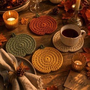 Peut inclure: Trois dessous de verre en crochet en forme de citrouille, de couleur orange, vert olive et jaune moutarde. Chaque dessous de verre présente une petite tige verte sur le dessus et un motif en spirale texturé. Une tasse blanche est posée dessus.