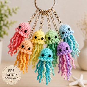 May include: A metal keyring featuring seven small, crocheted jellyfish charms in pink, orange, yellow, light green, turquoise, blue, and purple. Each jellyfish has black bead eyes, a stitched smile, and dangling curly tentacles. A circular label reads PDF PATTERN DOWNLOAD.
