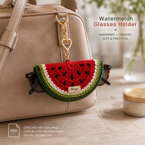 May include: A crochet watermelon glasses holder featuring a red, white, and green semi-circular design with black seed accents. It includes a gold-toned heart-shaped clasp and a small rectangular tag labeled Megz, attached to a beige handbag.