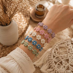 Può includere: Un set di cinque braccialetti a fiori realizzati a mano all'uncinetto in viola, rosa, bianco, blu e verde. Ogni braccialetto presenta una serie di piccoli motivi floreali interconnessi con una perlina color oro al centro di ogni fiore.