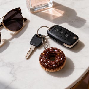 May include: A crocheted donut keychain featuring a dark brown top with colorful embroidered sprinkles and a tan bottom. The donut is attached to a silver metal keyring along with a black car key fob and a standard metal house key.